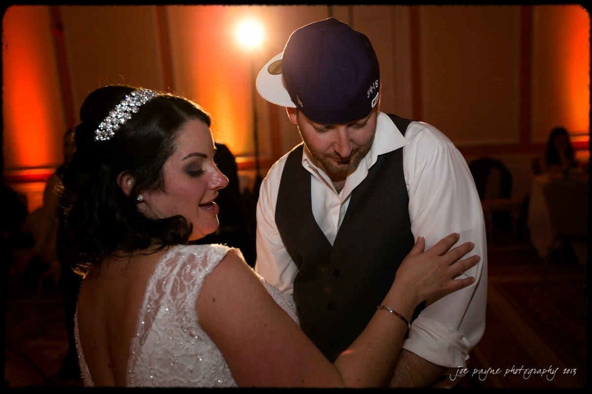 A couple is dancing at an event. The woman, wearing a white dress and jeweled headpiece, smiles as she looks at her partner. The man, in a vest and backwards cap, gently places his hand on her arm. The background is warmly lit and festive.