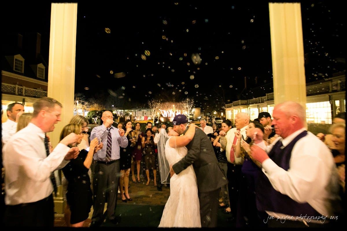 A bride and groom embrace under a night sky, surrounded by guests blowing bubbles. The scene is festive, with people clapping and celebrating in an outdoor setting. Lights illuminate the background, enhancing the joyful atmosphere.