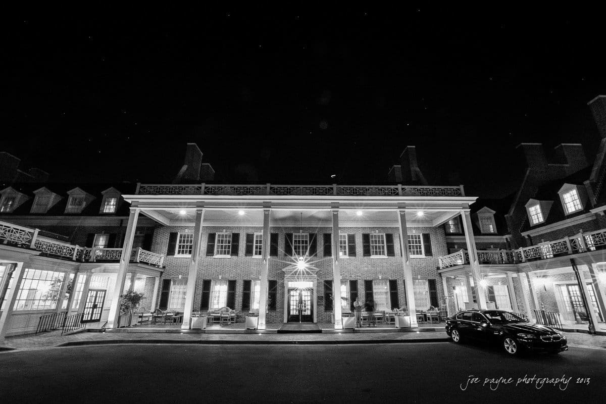 A black and white image of a grand, illuminated building with a large porch and columns at night. The entrance is lit, and a car is parked in front. Starry sky visible in the background.