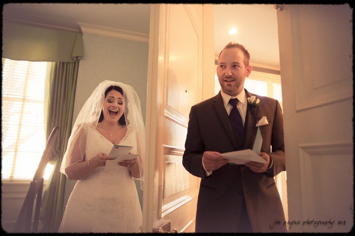A bride in a white dress and veil and a groom in a suit stand in a warmly lit room, both smiling while holding cards. The bride is near a window with drawn curtains, and the groom is near an open door.