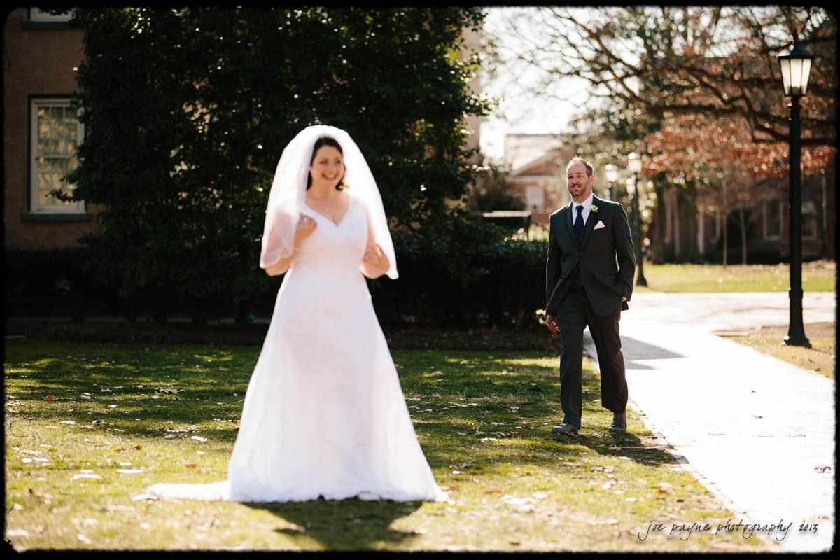A bride in a white gown and veil stands outdoors on grass, smiling. A groom in a dark suit and tie approaches from behind with a joyful expression. Sunlight filters through the trees, creating a warm atmosphere.