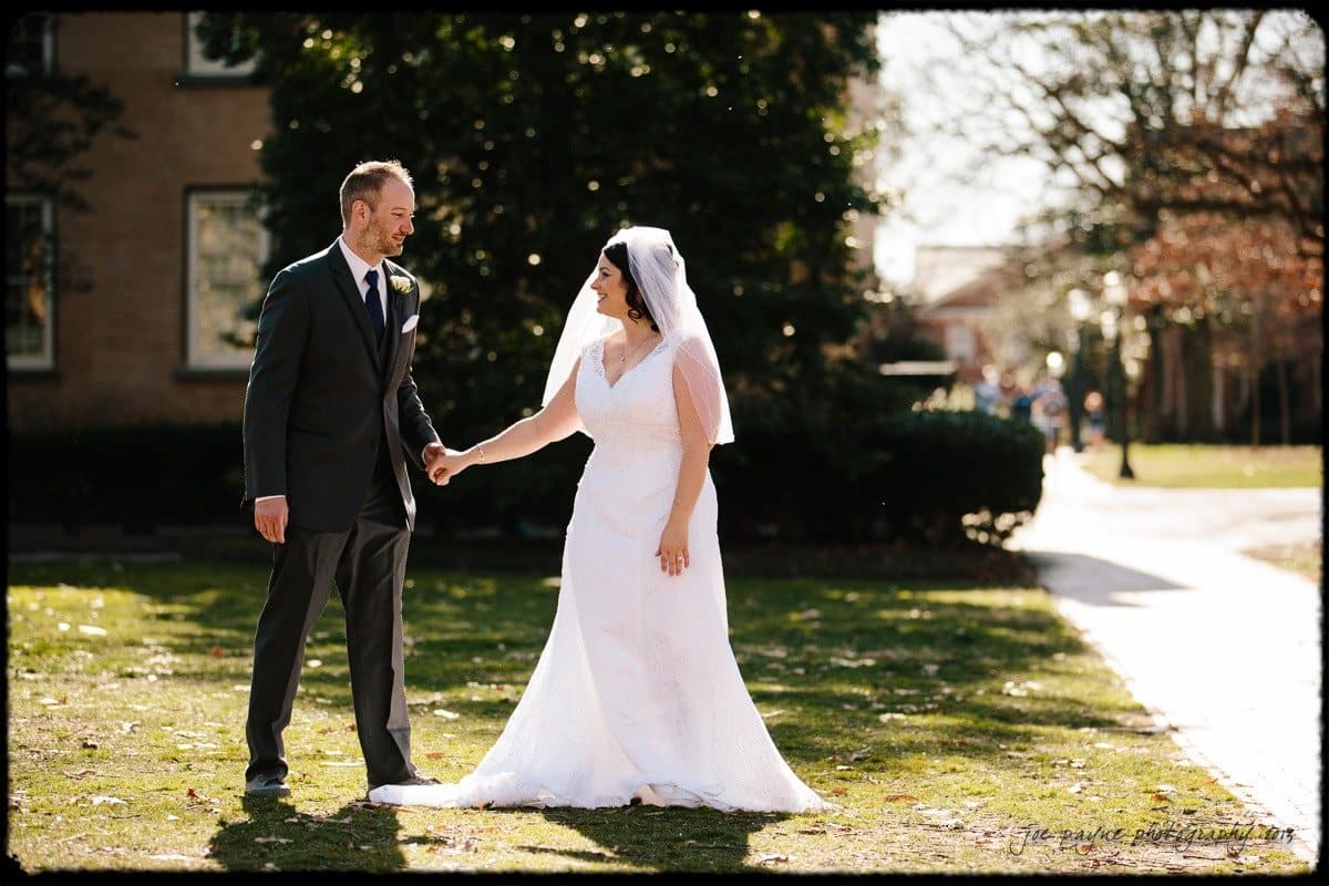 A bride in a white dress and veil holds hands with a groom in a dark suit and tie while standing on a sunlit lawn. Trees and buildings are in the background on a clear day.