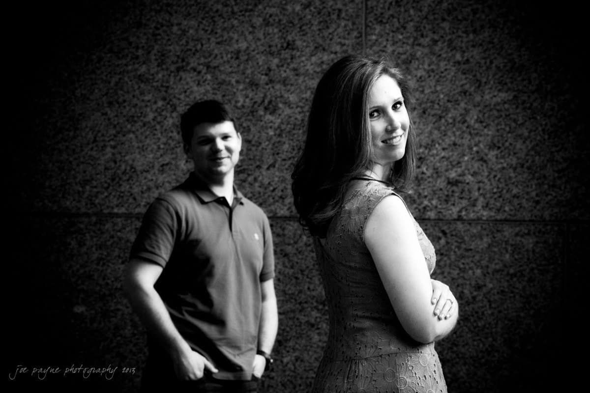 raleigh engagement photography ~ cassie and john A black-and-white photo of a woman smiling and looking over her shoulder at the camera, standing in front of a textured wall. In the background, a man stands with his hands in his pockets, also smiling.