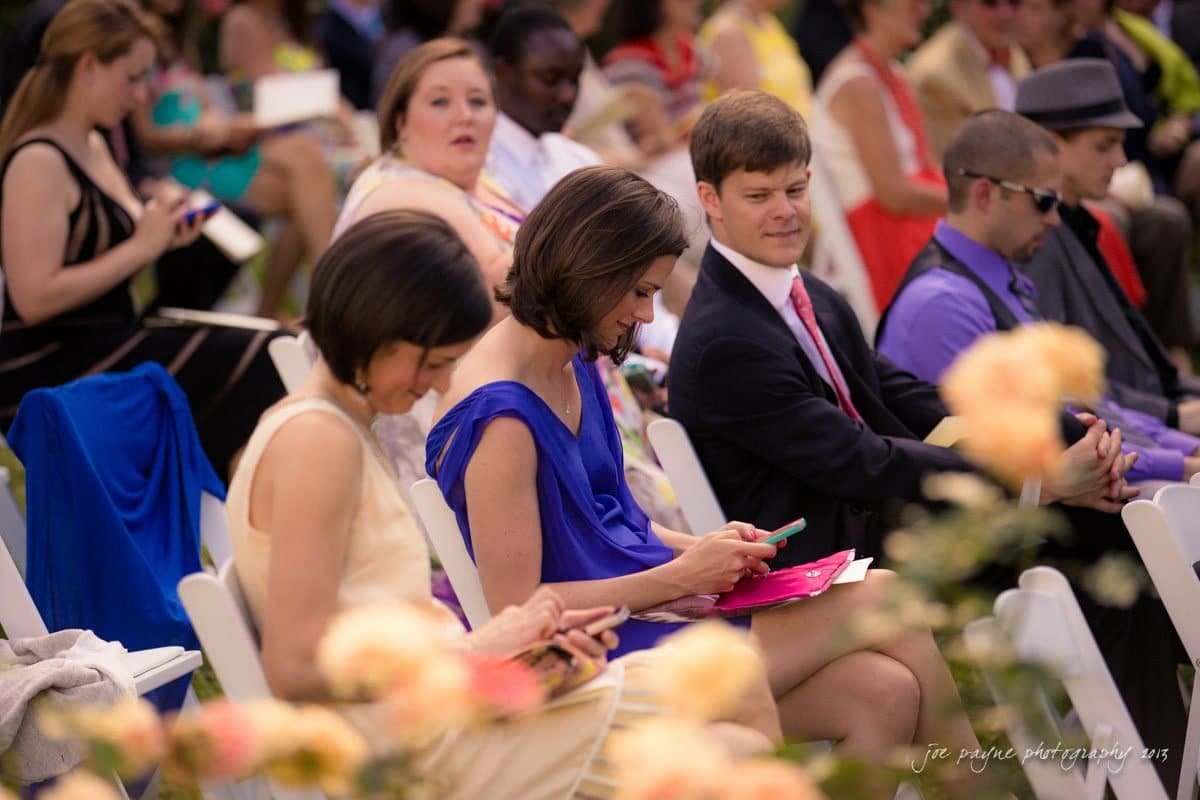Guests seated at an outdoor wedding. A woman in a blue dress uses her phone, while others engage in conversation. The foreground is blurred with flowers, and people in formal attire are in the background.