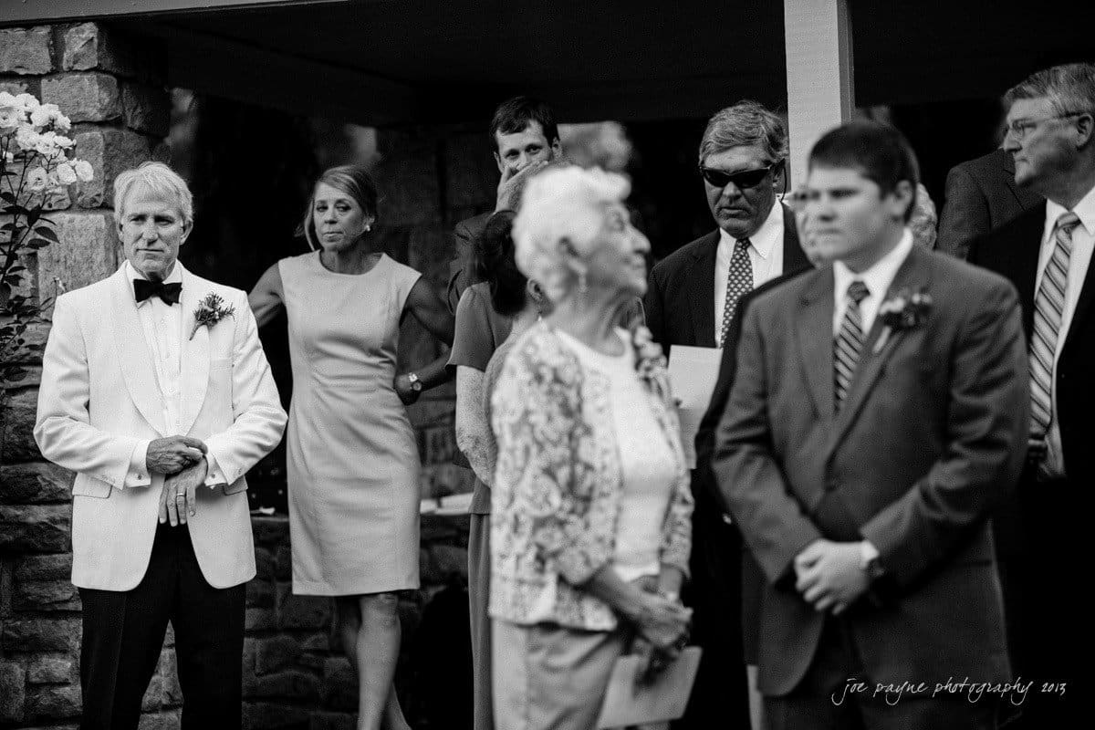A black and white photo of a formal gathering. A man in a white jacket with a bow tie stands on the left, surrounded by attendees. A woman in a dress is in the center, looking ahead, with others in suits and dresses around her.
