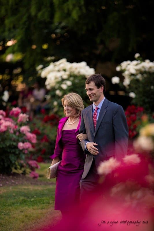 A woman in a purple dress and a man in a suit walk arm in arm through a garden filled with vibrant roses. The background is a lush green, complementing the colorful blossoms.
