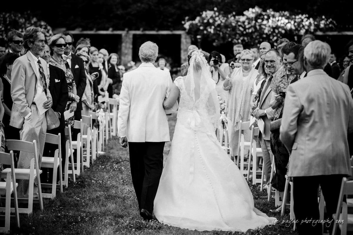 A bride and an older man walk down an outdoor aisle lined with seated guests on either side. The scene is set in a garden, and the guests, dressed in formal attire, are turned towards the couple. The photo is in black and white.