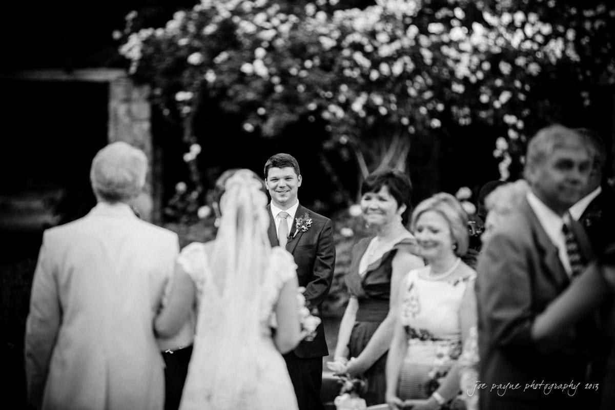 A black and white photo of a wedding ceremony. The bride is walking down the aisle towards the groom, who is smiling. Guests stand on either side, watching. The background is decorated with flowers.