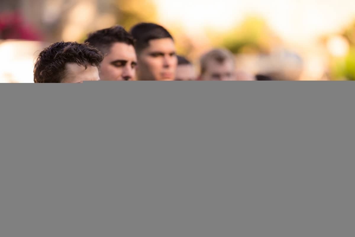 A group of people standing outdoors, their attention focused forward. The image is partially obscured, showing only the top portion of their heads. The background is blurred with hints of greenery and sunlight.