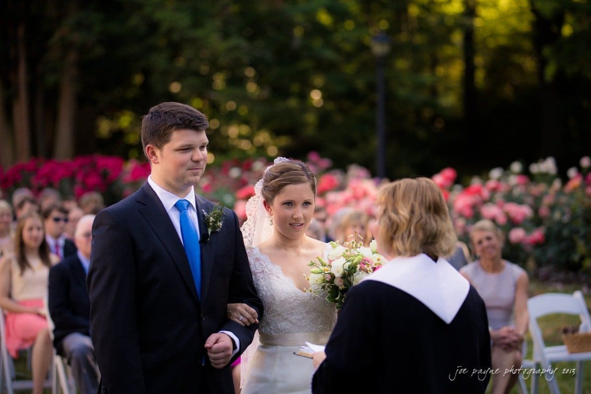 A couple stands together at an outdoor wedding ceremony. The groom is in a black suit and blue tie, while the bride wears a white dress and holds a bouquet. A person officiates the ceremony, with guests seated in the background.