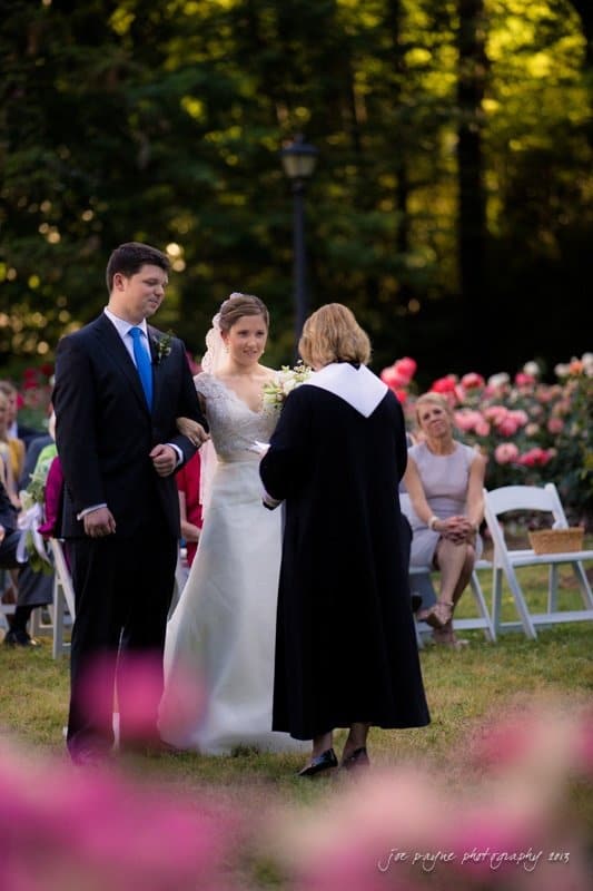 A bride and groom stand facing a minister during an outdoor wedding ceremony. The bride holds a small bouquet, and guests are seated in the background among vibrant flowers. The setting is serene with green trees and pink flowers in the foreground.