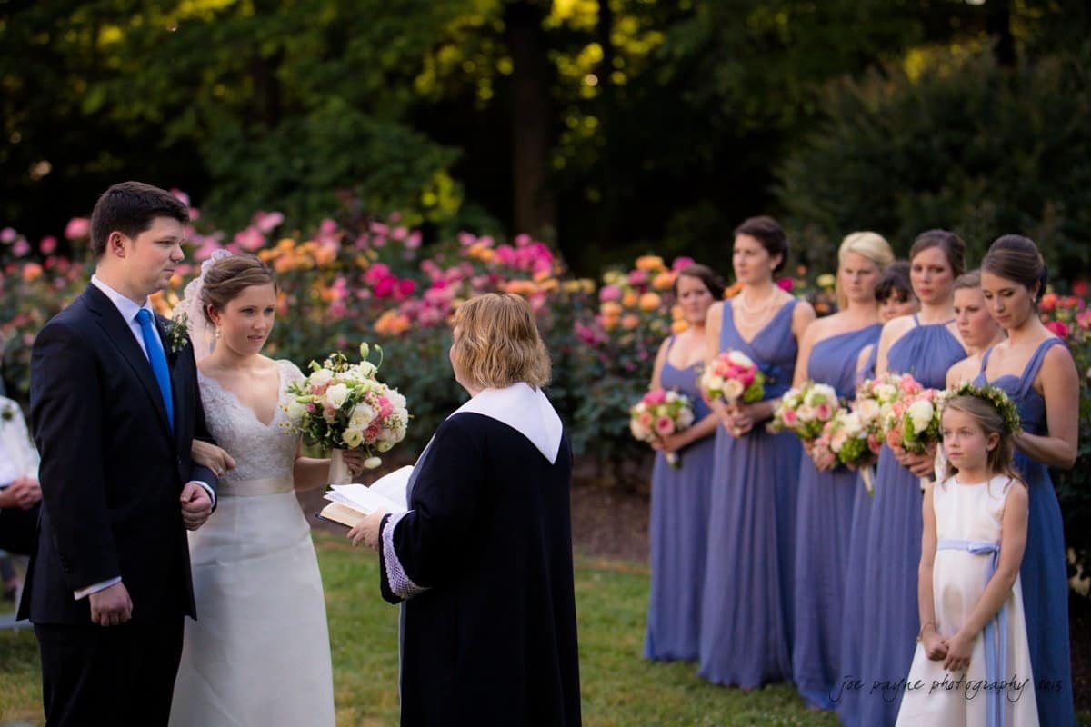 A bride and groom stand together during an outdoor wedding ceremony. The officiant reads from a book, and bridesmaids in lavender dresses hold bouquets in the background. A young flower girl stands to the side. The scene is surrounded by vibrant flowers.