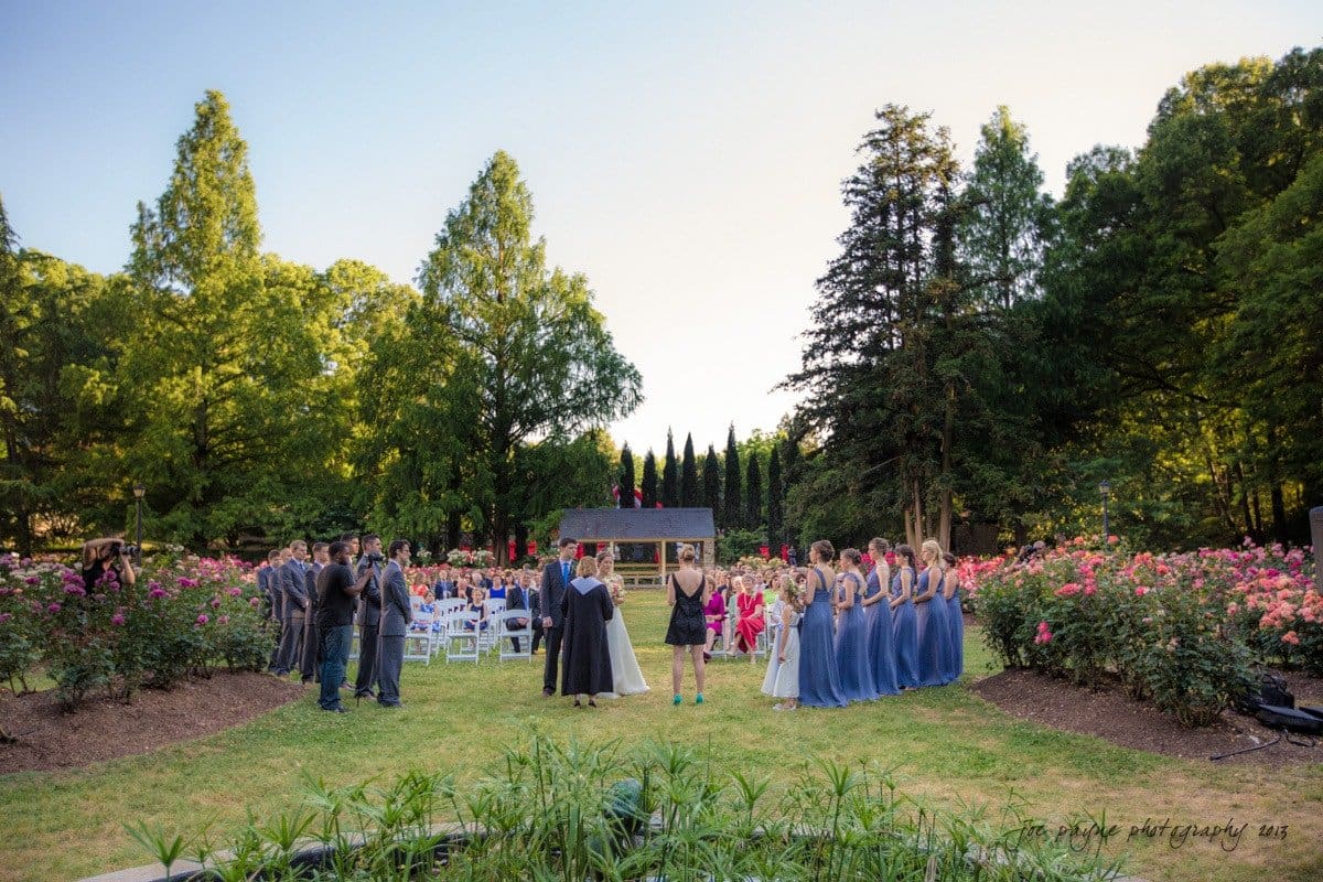 A wedding ceremony takes place outdoors in a garden filled with blooming flowers and surrounded by tall trees. The bride and groom stand at the altar, with bridesmaids in blue dresses and groomsmen in dark suits nearby. Guests are seated in white chairs.