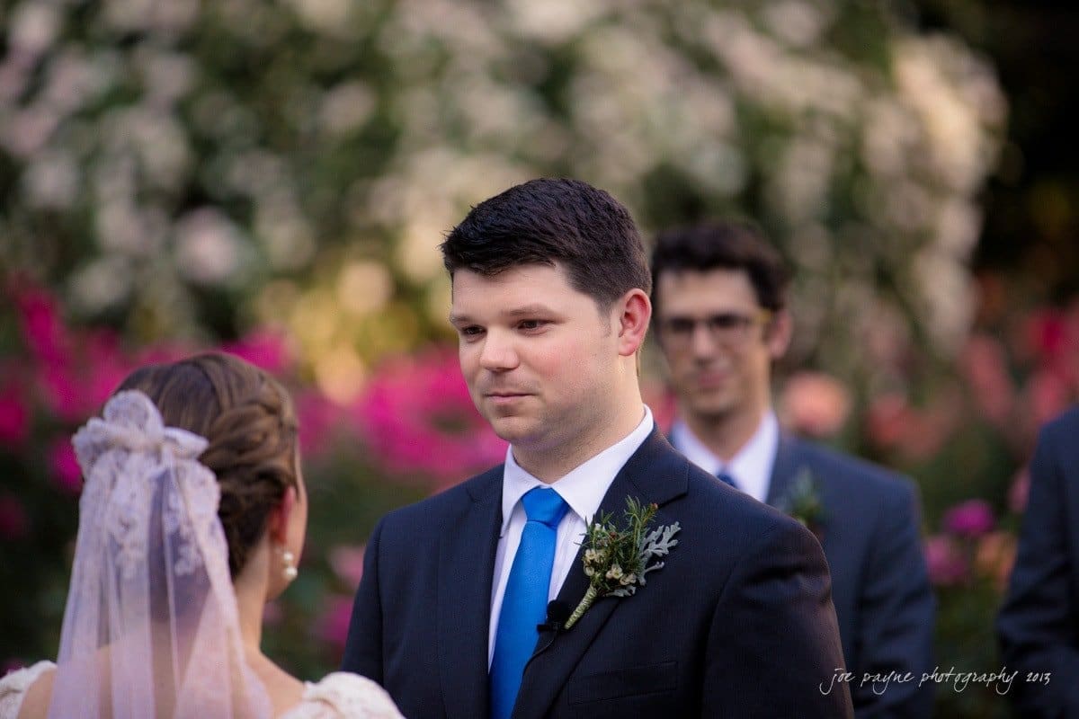 A groom in a blue tie, wearing a dark suit with a boutonniere, stands facing a bride in a wedding dress with a veil. Blurred greenery and a person in a blue tie can be seen in the background.