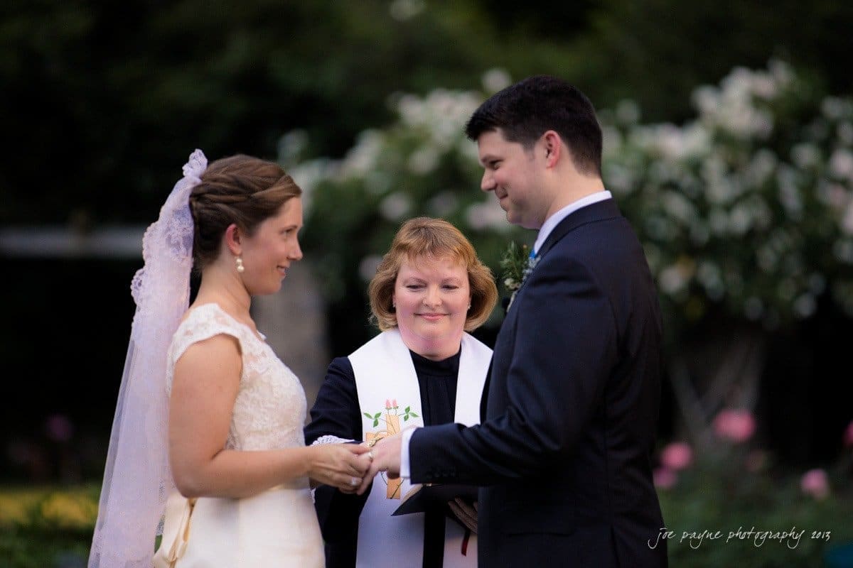 A bride and groom stand facing each other, holding hands during a wedding ceremony. A female officiant in a robe with a white stole stands between them, guiding the vows. The background is filled with greenery and soft-focus flowers.