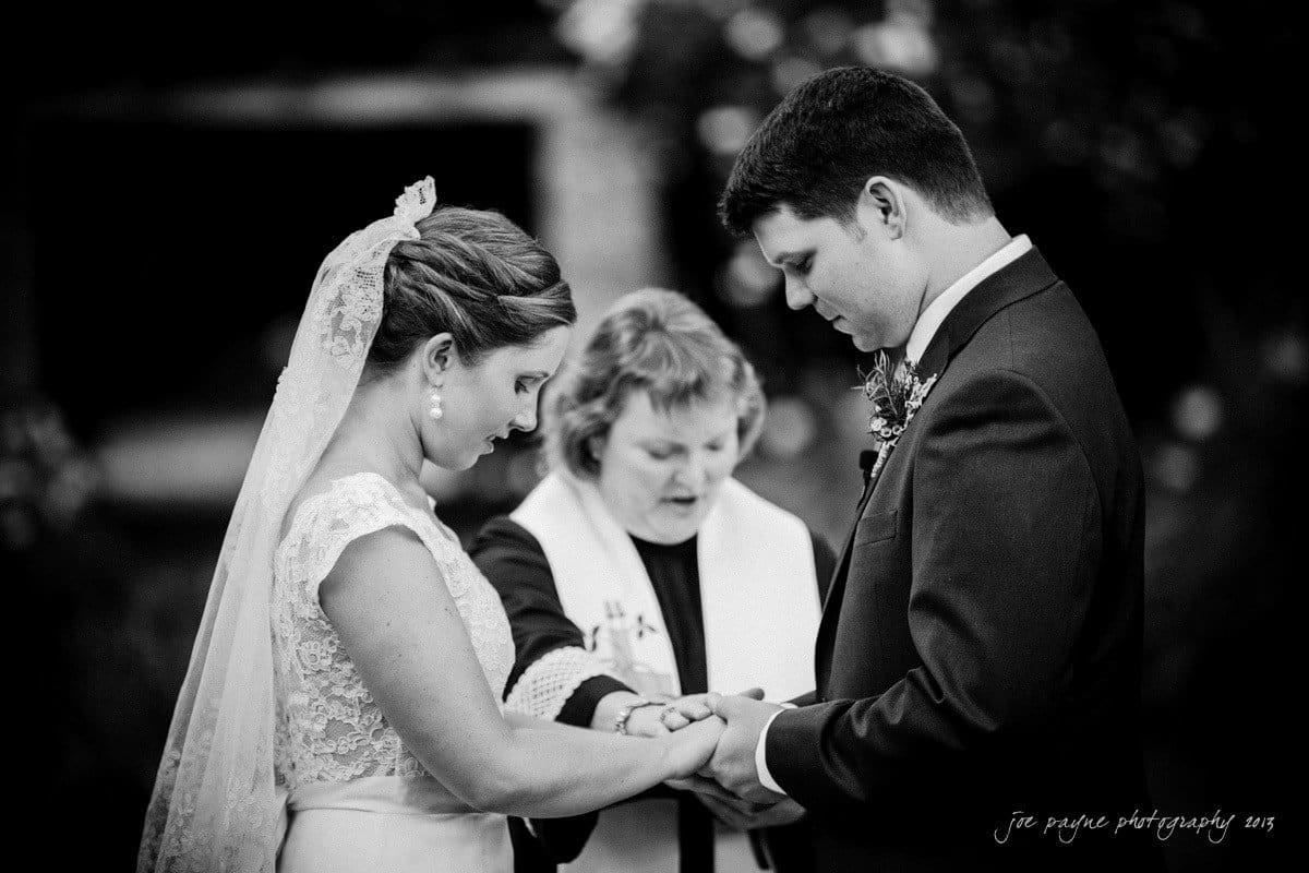 A bride and groom hold hands during an outdoor wedding ceremony, with an officiant standing between them. The bride wears a lace dress and veil, while the groom is in a dark suit. The scene is in black and white.