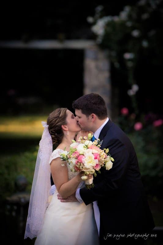 A bride and groom share a kiss outdoors, surrounded by greenery. The bride holds a bouquet of pink and white flowers, and the groom wears a dark suit. The background is softly blurred, highlighting the couples embrace.