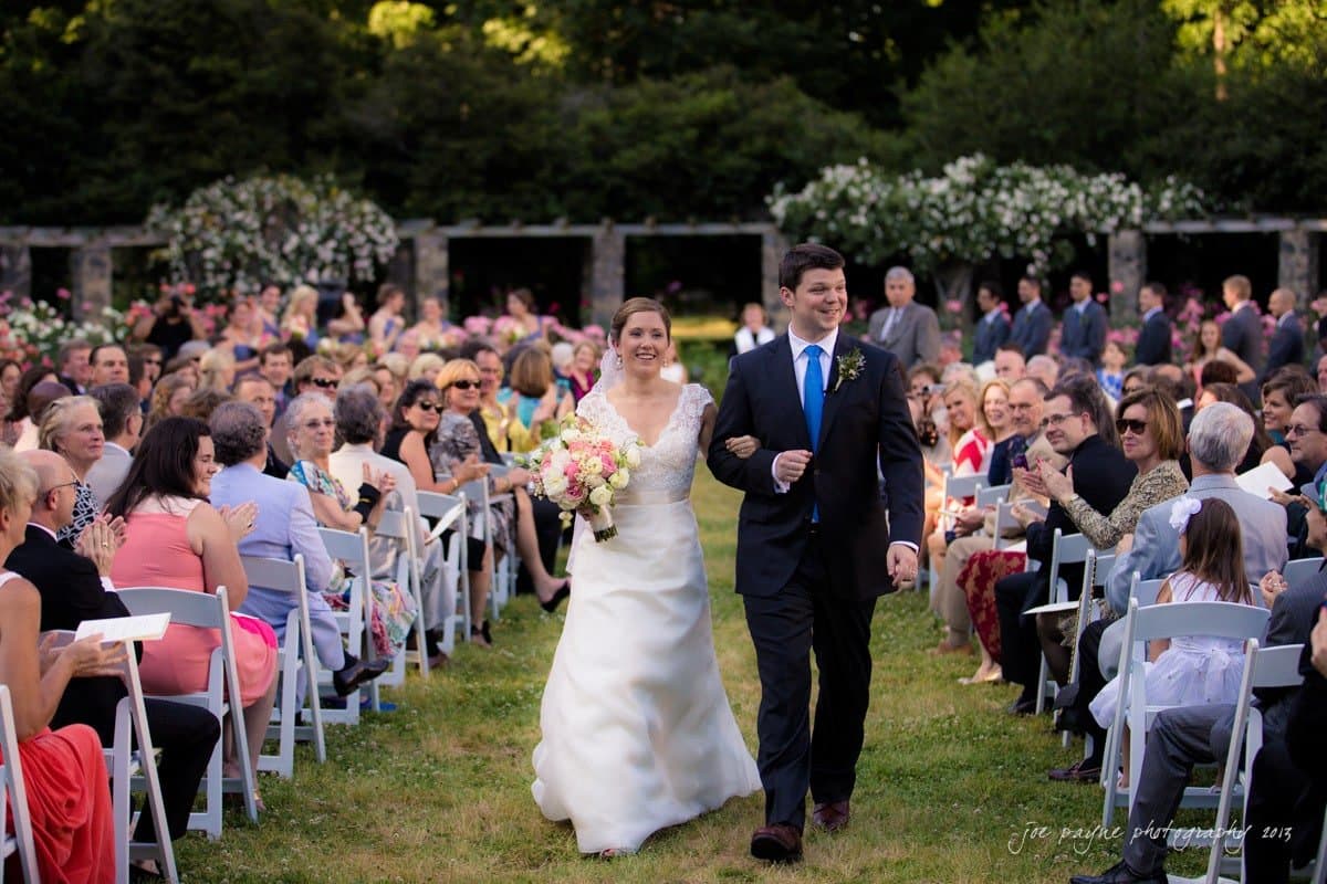 A bride and groom smile as they walk hand in hand down an aisle outdoors. The bride holds a bouquet of flowers, and they are surrounded by seated guests. The setting appears to be a garden or park with greenery and floral decorations.