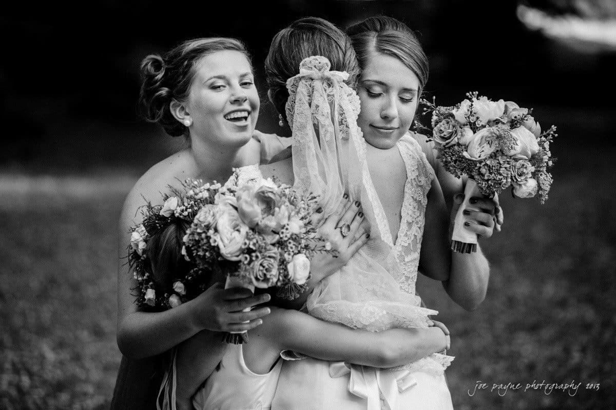 A black and white photo of a joyful wedding moment with four women embracing. They hold bouquets of flowers, with one woman wearing a wedding veil. The scene exudes warmth and celebration.