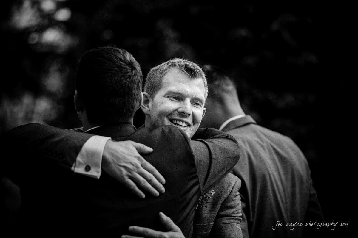 Two men in suits are embracing at an outdoor event. The man facing forward smiles brightly, while the other faces away from the camera. There are blurred onlookers in the background. The atmosphere appears warm and celebratory.