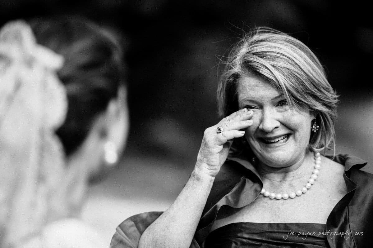 A black and white image of a woman wiping tears from her eyes while smiling. She wears a pearl necklace and an elegant dress. The focus is on her expressive face as she looks at another person in the foreground, whose back is to the camera.