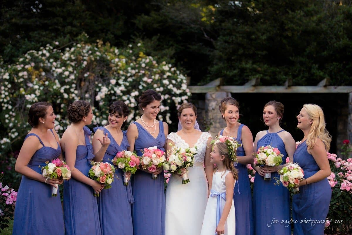 A bride stands smiling in a garden surrounded by seven bridesmaids and a young flower girl. The bridesmaids wear blue dresses, each holding a bouquet. The flower girl wears a white dress with a floral crown. Flowers and greenery are in the background.