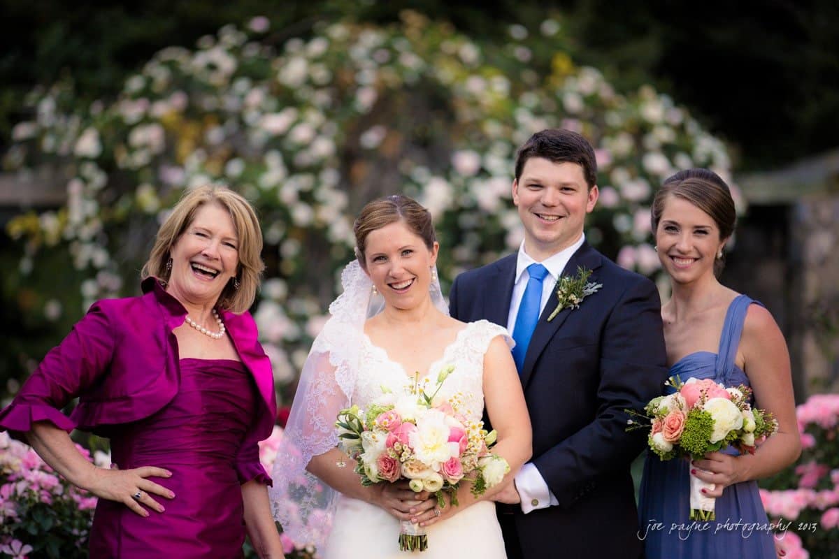 Four people smile at a wedding, with greenery and flowers in the background. The bride holds a bouquet and stands beside a groom in a suit. Two women, one in a purple outfit and another in blue, stand on either side, also holding flowers.