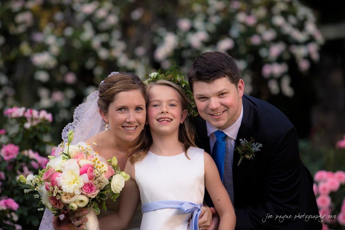 A bride and groom smile with a young girl between them. The bride holds a bouquet of pink and white flowers, and the girl wears a floral headpiece and a white dress with a blue ribbon. They are surrounded by blooming pink roses.