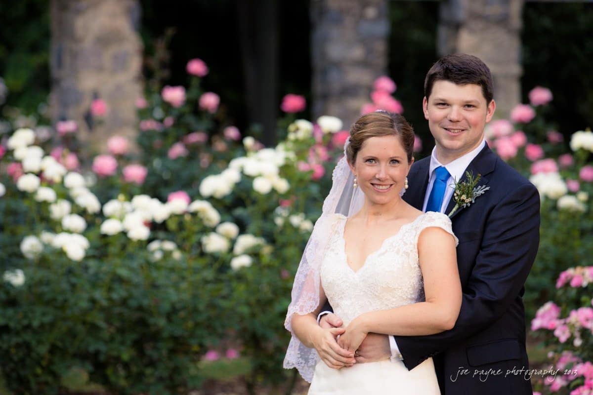 A bride and groom pose for a wedding photo in a garden. The bride wears a white gown and veil, holding a bouquet, while the groom in a dark suit and blue tie embraces her. They stand in front of blooming pink and white roses.