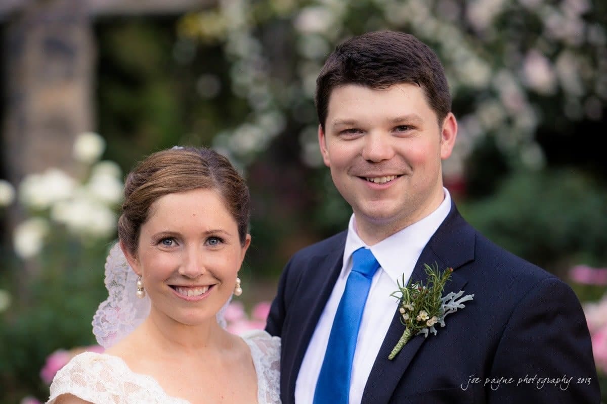 A bride and groom smile at the camera in an outdoor setting. The bride is wearing a white lace dress and veil, and the groom is in a dark suit with a blue tie. Flowers and greenery are in the background.