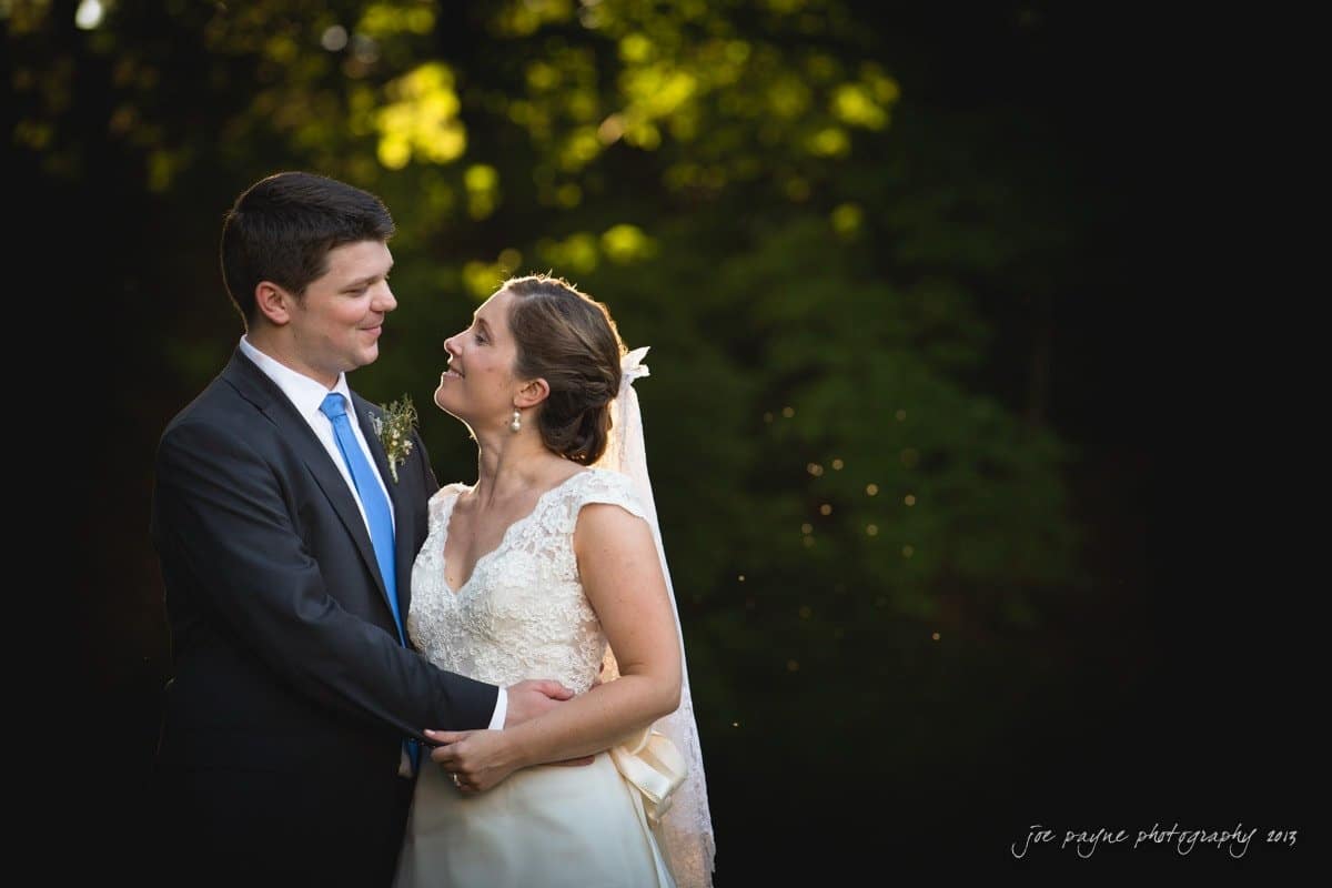 A bride and groom stand together outdoors, surrounded by greenery. The bride, wearing a white gown and veil, smiles at the groom, who is in a dark suit and blue tie. Sunlight filters through the trees, creating a warm, romantic ambiance.