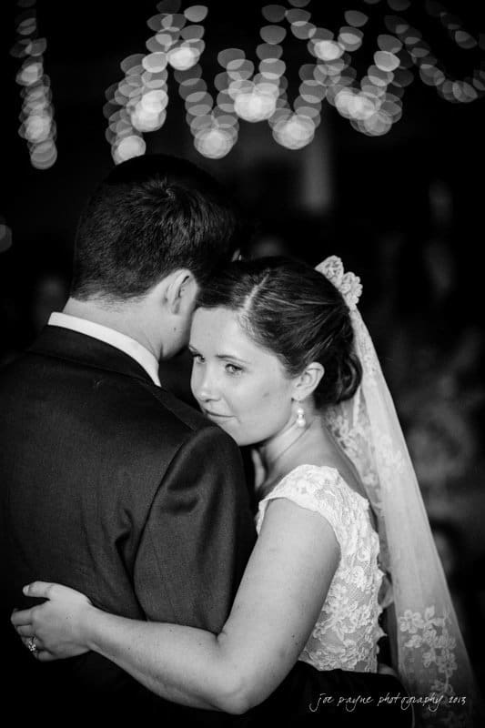 A couple embraces during a dance. The woman, in a lace gown with a veil, looks towards the camera while resting her head on the mans shoulder. Twinkling lights are blurred in the background, creating a dreamy atmosphere.