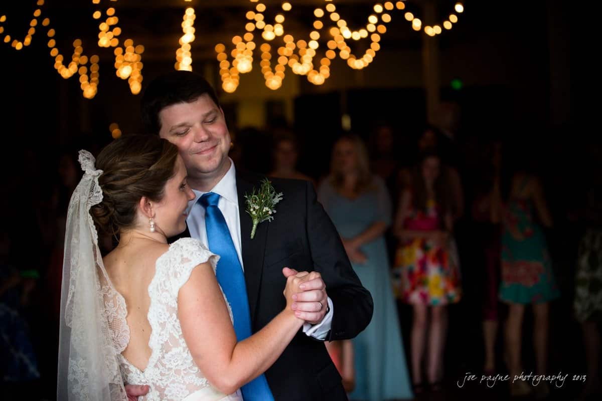 A bride and groom dance together at their wedding, smiling happily under strings of warm, glowing lights. The bride wears a lace gown and veil, and the groom is in a suit with a blue tie. Guests can be seen in the softly focused background.