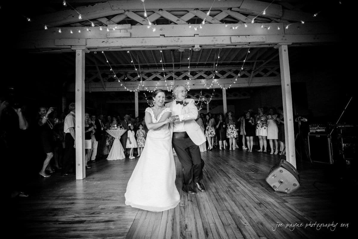 A couple dances joyfully in the center of a rustic hall decorated with string lights. The bride wears a flowing gown, and the groom is in a light suit. Guests watch and smile in the dimly lit background.