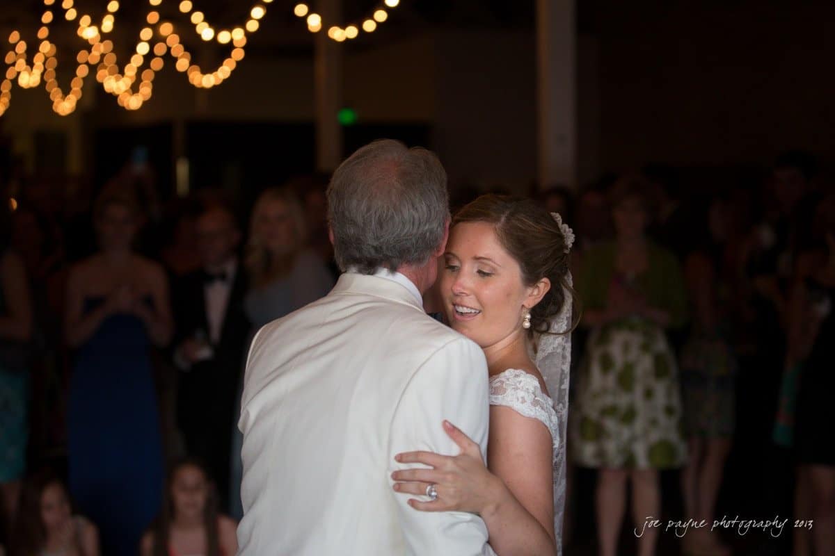 A bride and groom share a dance at their wedding reception. The bride is smiling, wearing a white dress and veil, while the groom is in a white suit. String lights hang above, and guests are visible in the softly lit background.