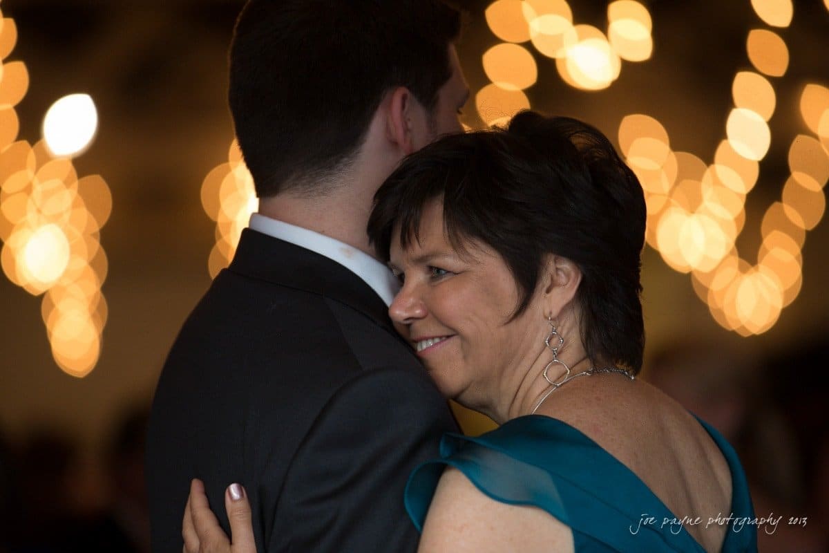 A woman in a teal dress smiles while dancing with a man in a suit. Warm, glowing lights create a soft, festive background ambiance.