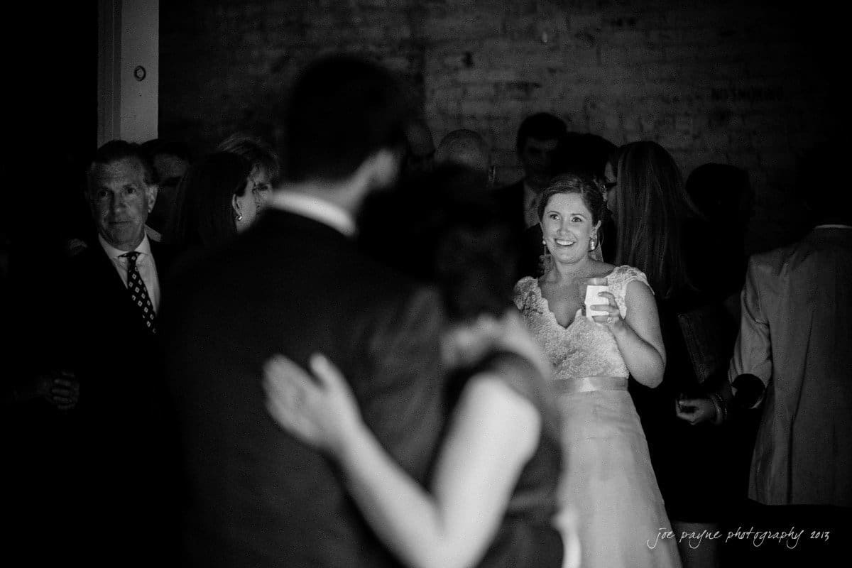A bride smiles while holding a cup, watching a dancing couple in a dimly lit room filled with guests.