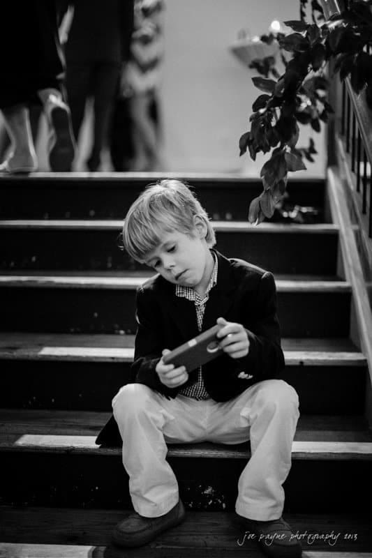A young boy sits on stairs, absorbed in a smartphone. He wears a suit jacket and white pants. The black-and-white photo captures a calm moment; leafy plants are in the background.