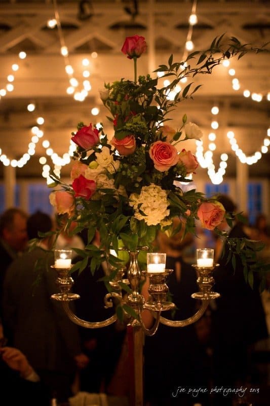 Tall floral centerpiece with pink and white roses, greenery, and lit candles on a candelabra. Soft, warm lighting with string lights in the background. Elegant event or celebration setting.