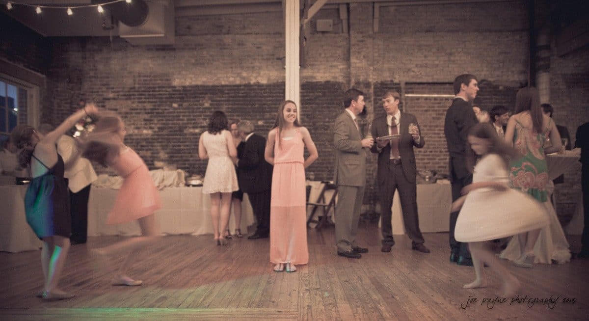 A girl in a pink dress stands smiling in the center of a rustic room with exposed brick walls, while children in motion blur around her. People in formal attire gather near a buffet table in the background.