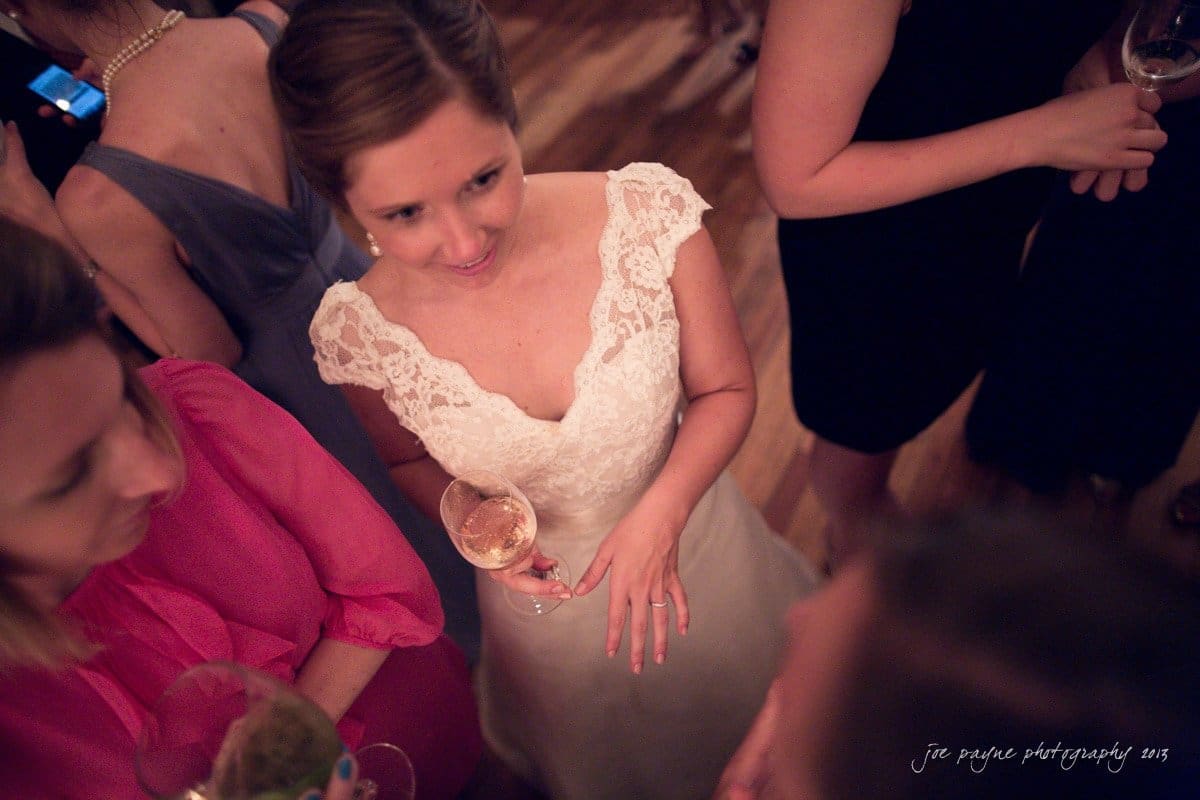A bride in a white lace dress holds a glass of wine, standing among guests at a gathering. People around her are engaged in conversation, and the atmosphere appears lively and festive.