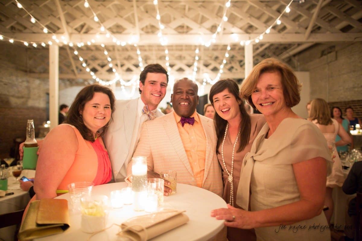 A group of five people, dressed in formal attire, smile together at a round table. The setting is a warmly lit indoor venue with string lights and exposed brick walls, suggesting a festive event or celebration.