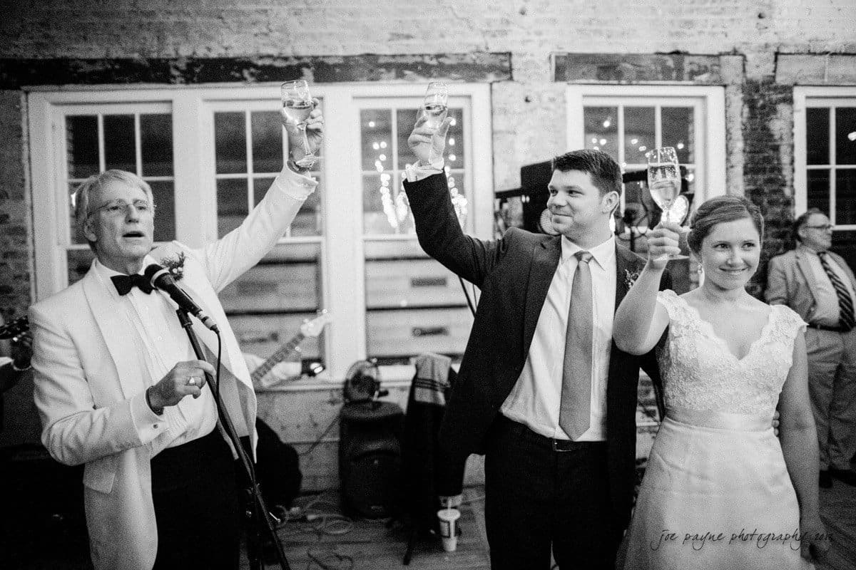 A black and white photo of three people standing and raising their glasses for a toast at a wedding. The man on the left is speaking into a microphone. The group is in front of a brick wall with large windows.