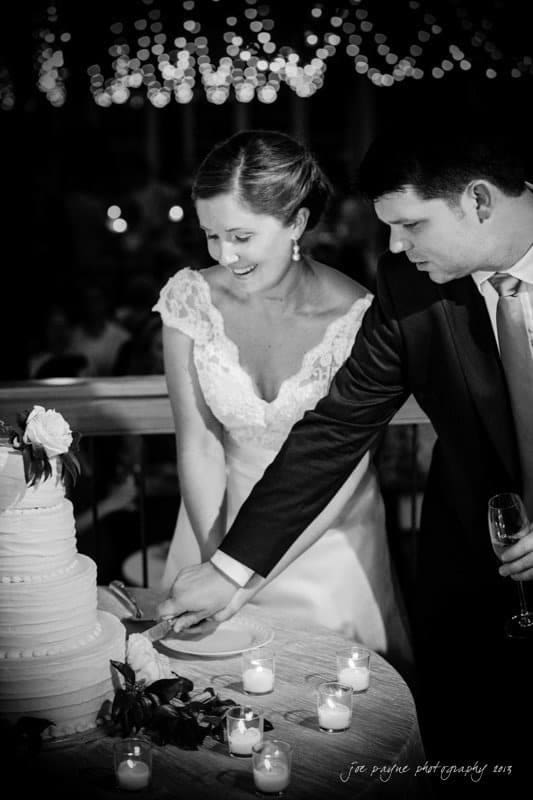 A bride and groom are cutting a three-tiered wedding cake together. The bride is smiling, and the groom is focused on the cake. They are surrounded by small lit candles, with string lights visible in the background.