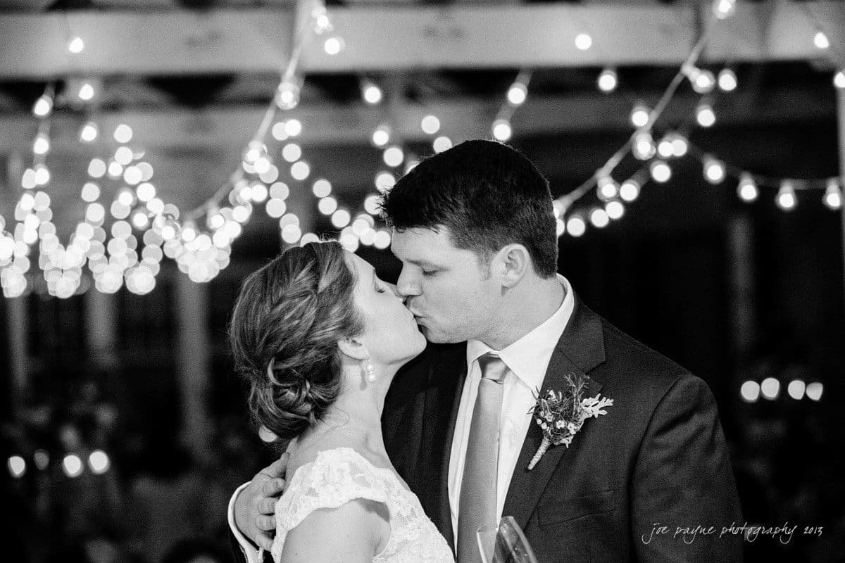 A couple is sharing a kiss at their wedding under a canopy of string lights. The bride is in a lace dress with her hair up, and the groom is in a suit with a floral boutonniere. The background is softly blurred, emphasizing the intimate moment.