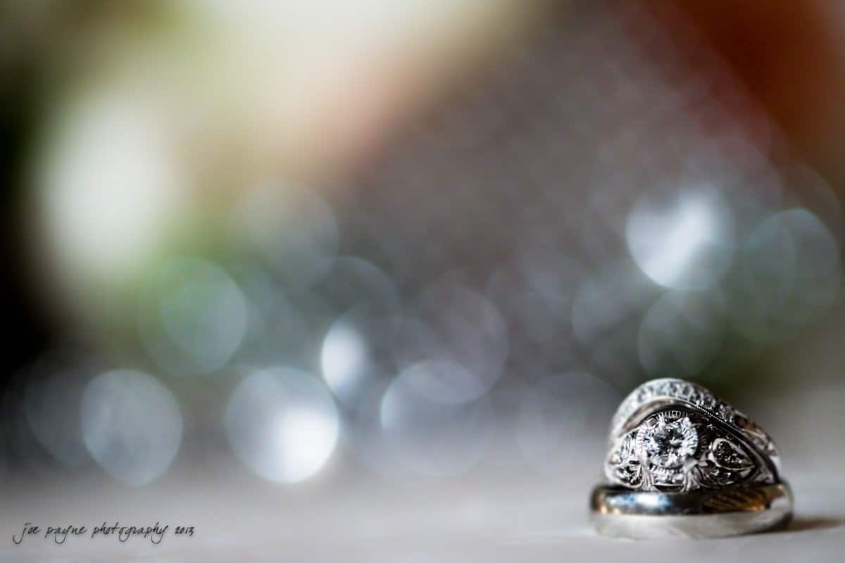 Close-up of two elegant silver rings on a soft surface, with one featuring an ornate design and a prominent round diamond. The background is softly blurred, creating a bokeh effect with circular light patterns.