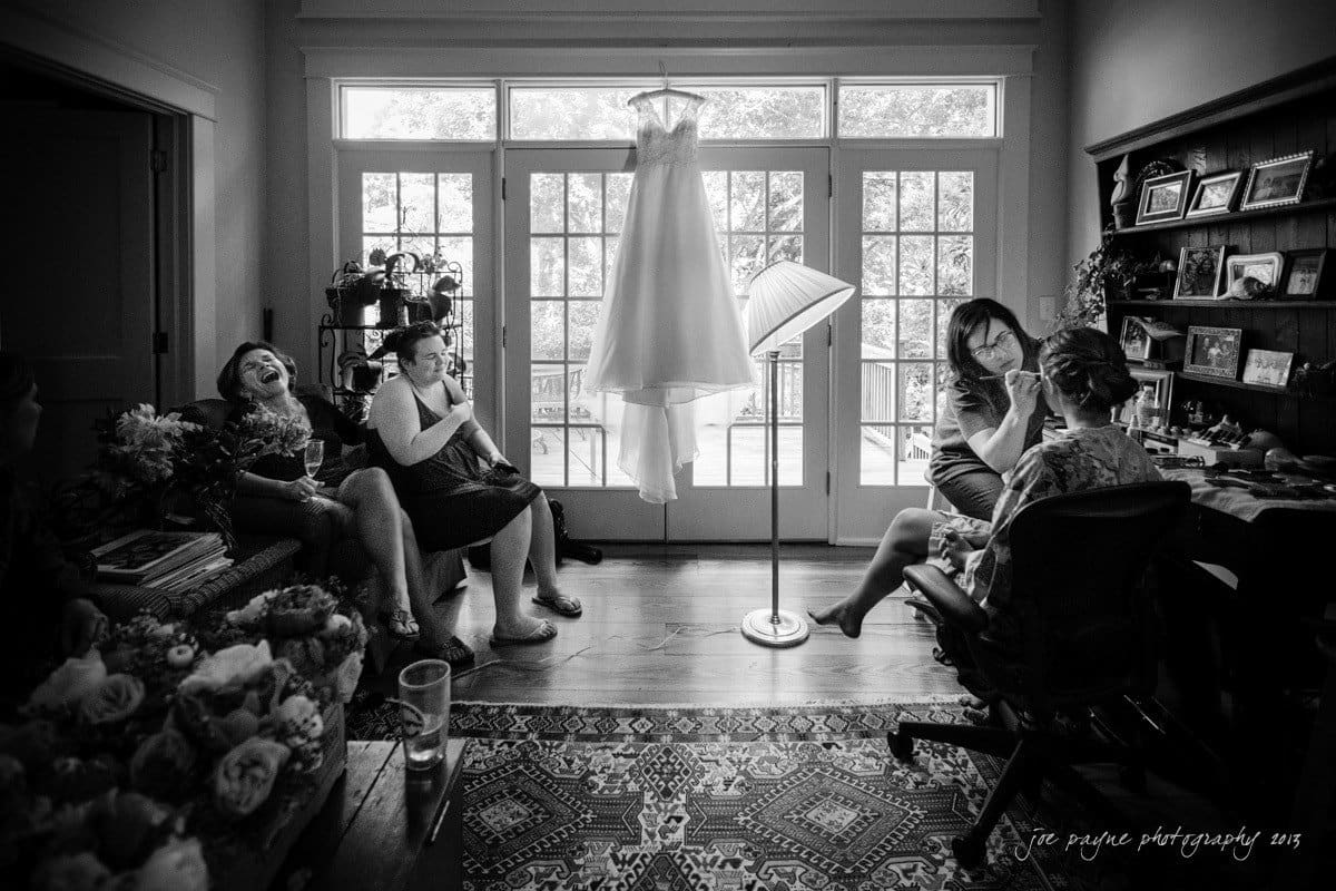 A bride gets ready in a room with a long white wedding dress hanging by a large window. Three women sit nearby, smiling and chatting. The room is warmly lit, with flowers and a decorative rug adding to the cozy atmosphere.