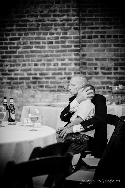 A black and white photo of a man seated at a table, cradling a sleeping child on his shoulder. The table holds bottles, glasses, and a brick wall is in the background. The setting appears calm and intimate.