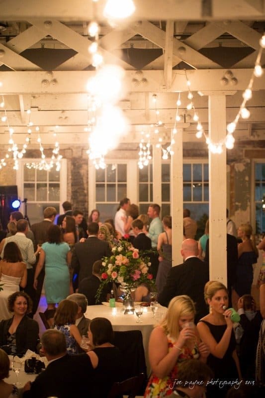 A lively wedding reception with guests dancing under string lights in a rustic venue. Tables are adorned with floral centerpieces, and people are socializing, creating a warm and festive atmosphere.