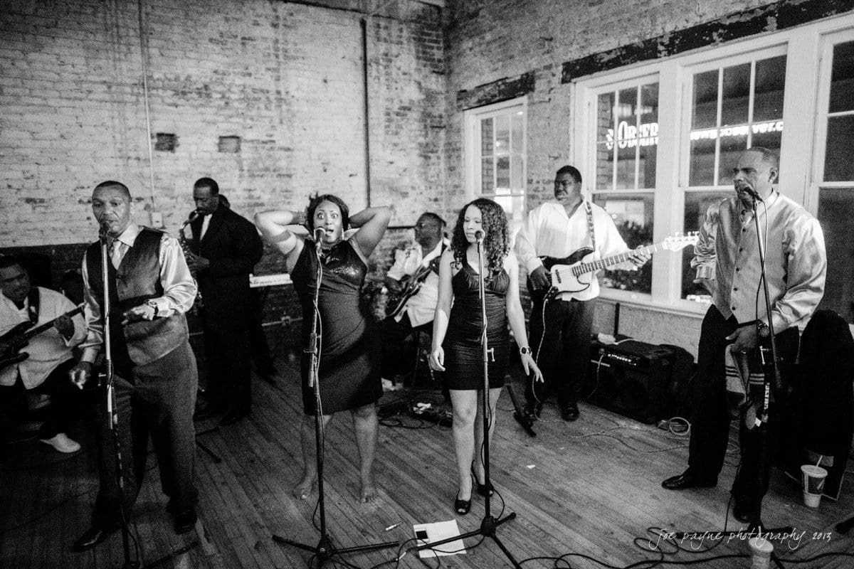 A black and white photo of a band performing indoors. Two women at the front sing into microphones, with male musicians playing guitar and keyboard in the background. The setting has exposed brick walls and wooden floors, creating a vintage atmosphere.